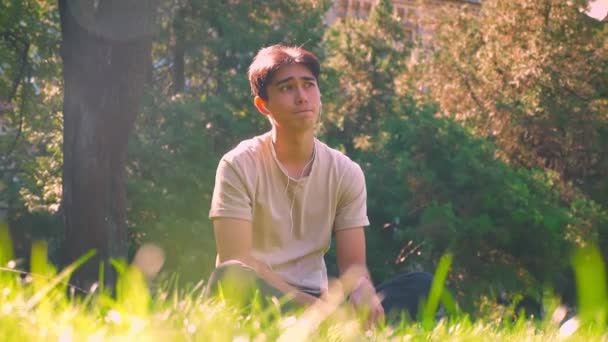 Dreaming asian boy is hanging out alone in park, sitting on the grass in sunlights, fresh spring atmosphere — Stock Video