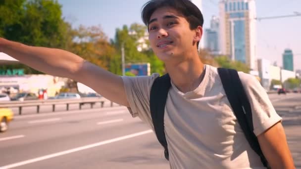 Nice asian man in backpack is hitchhiking with great hopeful face, vista urbana na estrada, dia ensolarado — Vídeo de Stock