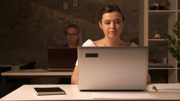 Dos mujeres caucásicas de trabajo está sentado en la oficina de ladrillo marrón y el uso de sus computadoras portátiles en el escritorio blanco interior — Vídeos de Stock