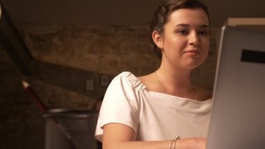 Utangaç gülen kafkas kadın dizüstü bilgisayarda çalışan ve kapalı, kahverengi tuğla duvar otururken doğruca kameraya bakıyor