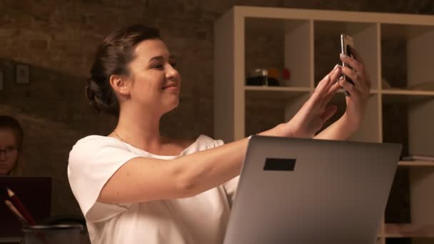 Niedliche kaukasische Geschäftsfrau macht Selfie, während sie neben einer anderen erwachenden Frau in der Nähe von Laptop im braunen Studio sitzt — Stockvideo