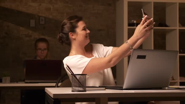 Grande femmina caucasica sta prendendo selfie sul suo smartphone mentre seduto in ufficio con la sua collega, scrivanie separate — Video Stock