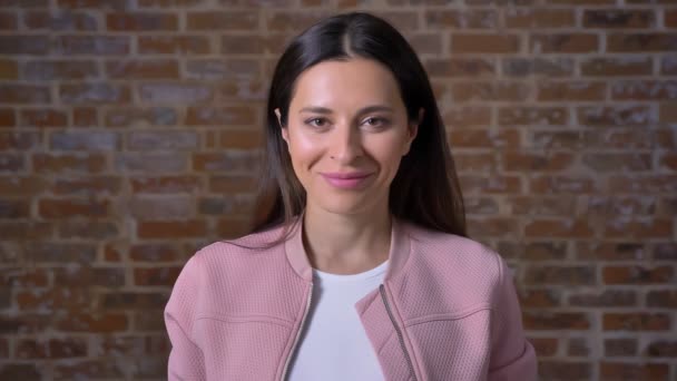 Disfrutable chica caucásica está de pie delante de la pared de ladrillo y sonriendo sinceramente a la cámara, look casual, ladrillo estudio de tiro — Vídeo de stock