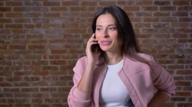 Gülen muhteşem beyaz kadın koyu uzun saçlı telefon üzerinden duygusal olarak tuğla arka plan ayakta konuşuyor