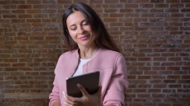 Glücklich awesome kaukasischen Weibchen zeigt grünen Bildschirm auf ihrem Tablet, zeigt mit einem Finger darauf und steht auf Backstein-Hintergrund allein — Stockvideo