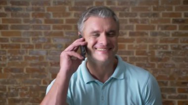 Duygusal olgun adam telefonla konuşmak ve kamera ile tuğla duvarın arkasında ayakta sohbetteki çıkıldı, ciddi görünümlü