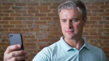 Ciddi üst düzey erkek onun cep telefonu tutarak ve süre ayakta ve biraz etrafında, brick kırmızı stüdyo hareketli kameraya bakarak