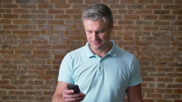 Schöner reifer kaukasischer Rüde scrollt konzentriert sein Handy, während er in blauem T-Shirt neben einer Ziegelwand steht — Stockvideo
