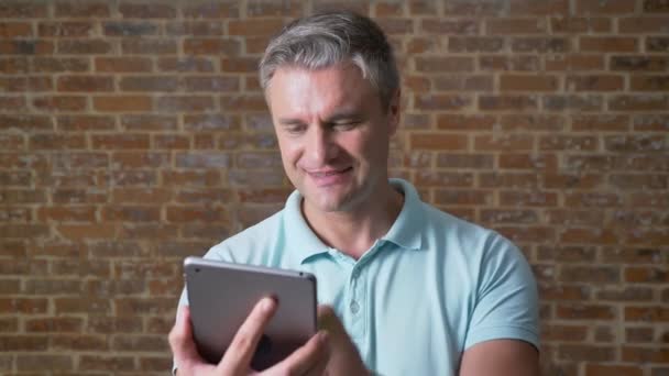 Eccitato maturo uomo caucasico con i capelli grigi sta mostrando schermo verde sul suo tablet e sorridente, tiro cluse-up, sfondo di mattoni — Video Stock