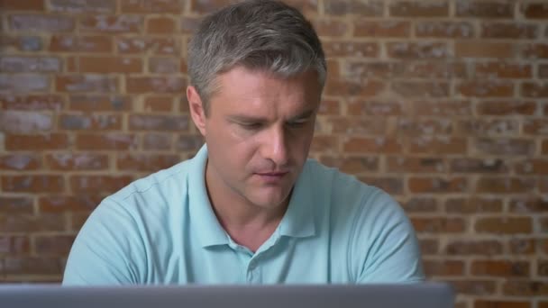 Beau caucasien homme d'affaires adulte est assis à côté de cet ordinateur portable et profiter de sa boisson de la tasse, isolé sur fond de brique — Video