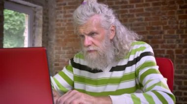 Komik üst düzey beyaz gri adam odaklı ve mutlu bir şekilde kameraya gülümseyen yüzünde modern tuğla ofisinde oturuyor ve onun laptop içinde yazarak