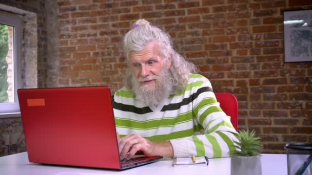 Agradable abuelo caucásico está utilizando la computadora roja y estirando su cuerpo para relajarse mientras está sentado en el estudio de ladrillo rojo — Vídeo de stock