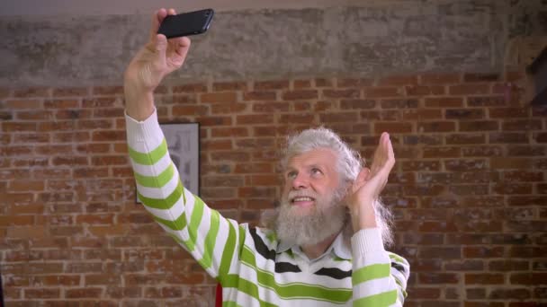 Mignon vieux caucasien barbu mâle agite à la caméra avant de son smartphone et souriant détendu, longs cheveux blancs, brique studio rouge — Video