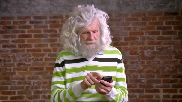 Schattige harige witte baard oude man is zijn telefoon staan en kijken naar camera met ontspannen lachende gezicht, permanent geïsoleerd in de buurt van muur schuiven — Stockvideo