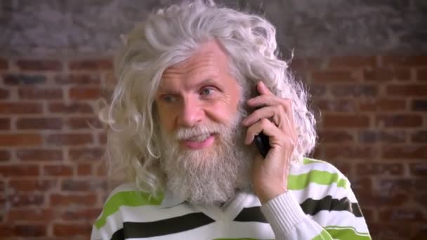 Calma conversazione per telefono di vecchio maschio caucasico con grande barba bianca e capelli ondulati, focalizzata o la sua chiamata e in piedi su sfondo di mattoni, moderna vista chill — Video Stock