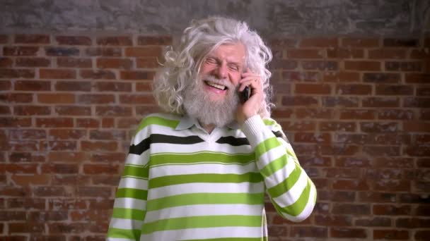 Amusant homme caucasien âgé aux cheveux blancs et à la barbe lourde, accroché à son téléphone et parlant avec un visage souriant, illustration de bonheur, fond de brique rouge — Video