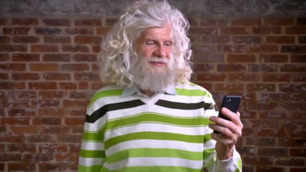 Крутой старший кавказский мужчина с большими белыми вьющимися волосами и красивой бородой проводит видеозвонок и спокойно разговаривает, стоя в современной кирпичной студии в одиночестве — стоковое видео