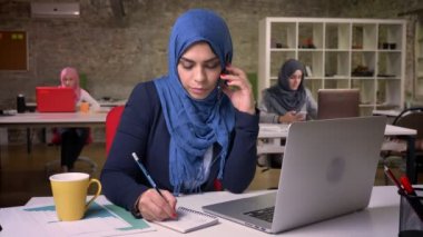 Clamly telefonu musun modern tuğla Office arkasında meslektaşları ile oturan arabaian kız mavi hijab ve defter, çeşitlilik günümüzde yazma