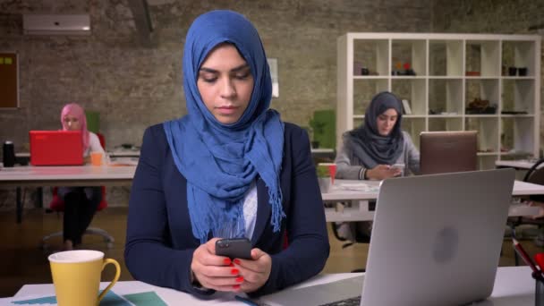Ernstes Gesicht einer amuslim Frau in blauem bezaubernden Hijab scrollt ihr Handy, während sie im modernen Studio sitzt, umgeben von anderen arbeitenden arabischen Mädchen — Stockvideo