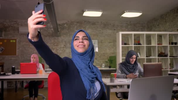 Heppy Arabská žena bere selfie na telefonu zatímco sedí v křesle v kanceláři cihel s dalšími muslimskými děvčaty, moderní podnikatelky — Stock video