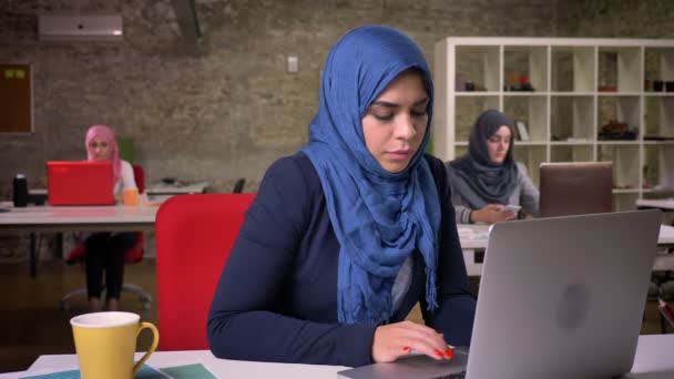 Зияющая арабская женщина в темно-синем хиджабе работает над своим ноутбуком, в то время как другие офисные женщины ias сидит позади, внутренняя иллюстрация — стоковое видео