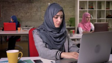 Doğruca kameraya, ışık kapalı, güzel modern mecaz seyir Ortadoğu kız oturma ve bilgisayar başında, çalışma hijab içinde neşeli bir gülümseme