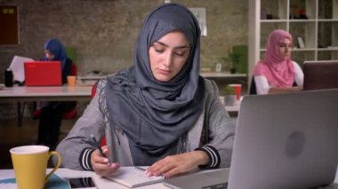 Hijab şirin Arap kadın notlar aşağı yazma ve kontrol onun laptop, çalışma vibes, meslektaşları arkasında, çeşitli modern yaşamı