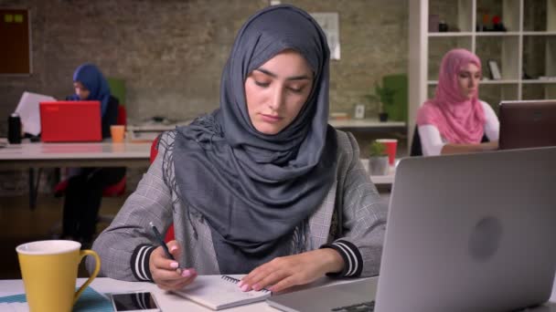 Милая арабская женщина в хиджабе записывает заметки и проверяет свой ноутбук, рабочие вибрации, коллег позади, разнообразная современная жизнь — стоковое видео