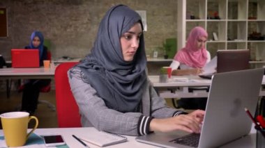 Modern arabic woman is typing on laptop, looking precisely at the screen, working mood, sitting at workplace, females in hijabs on background, brick office