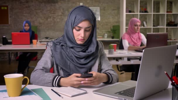 Koncentrált szép muszlim lány hidzsáb ül a munkahelyen, és ő mobiltelefon közelében vele laptop és más arab lányok háttér — Stock videók