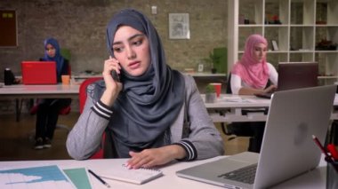 Hijab emin alışılmadık Müslüman kız ciddi konuşma telefonda vardır oturma, modern vibes, tuğla ofis, arka plan üzerinde Arap kadınlar