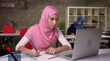 Güzel çalışma Arap Erkek pembe hijab ve İslami giyim notlar aşağı yazma ve onu laptop tam önünde oturma diğer Müslüman kadınlar, office vibes süre seyir