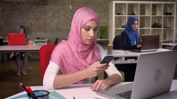 Mooie Arabische meisje in roze hijab is typen met op vinger een haar telefoon chill zittend op haar werk, collega's op de achtergrond, binnen licht groene bakstenen kantoor — Stockvideo