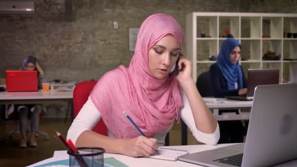 Hübsche Frau aus dem Mittleren Osten im pinkfarbenen Hijab spricht selbstbewusst und ruhig über ihr Smartphone, schreibt am Arbeitsplatz, nebenbei illustriert, arbeitet heutzutage — Stockvideo