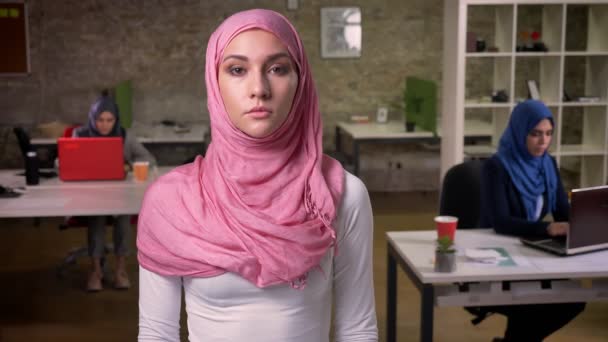 Bella donna araba è in piedi con calma in hijab rosa e guardando la fotocamera dritta mentre altre femmine sono sedute e lavorano in ufficio mattoni — Video Stock