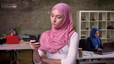 Pembe hijab giyen güzel Müslüman kız ayakta ve onun smartphone modern ofis içinde Orta Doğu kızlar arka plan üzerinde tutarak.