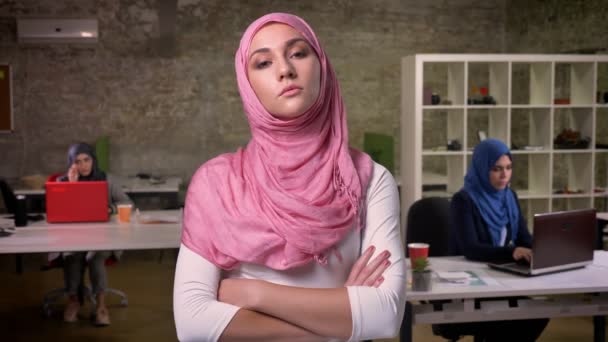 Gadis arab yang tenang dalam jilbab merah muda berdiri dengan jilbab merah mudanya di kantor bata dan melihat kamera dengan percaya diri, duduk gadis arab di latar belakang — Stok Video