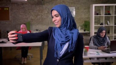 Keyifli Arap kadın gülümseyerek ve selfie mavi Hicap alarak ve düz ayakta frocess, kapalı, tuğla ofis odaklı