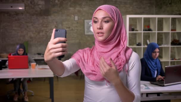 Aranyos arab nő a rózsaszín szép hidzsáb, amelynek videohívás állva tégla modern szobában, neki dolgozó kollégák közelében — Stock videók