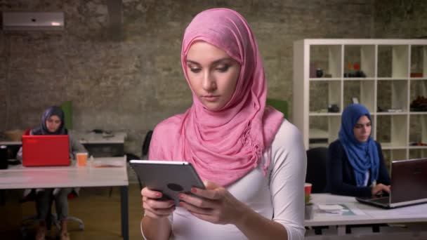 Concentrato arabo femmina sta digitando sul suo tablet mentre in piedi vicino ad altre ragazze arabe in mattoni ufficio moderno separato, illustrazione indoor — Video Stock