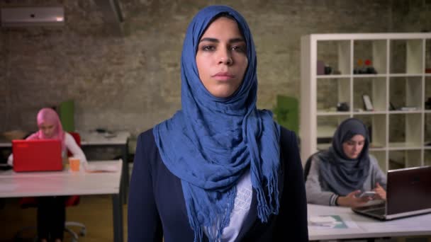 Wanita arab yang bangga dan percaya diri dalam jilbab biru tua melihat langsung ke kamera sambil berdiri diam dengan tangan disilangkan, kantor modern bata, gadis-gadis di latar belakang bekerja — Stok Video