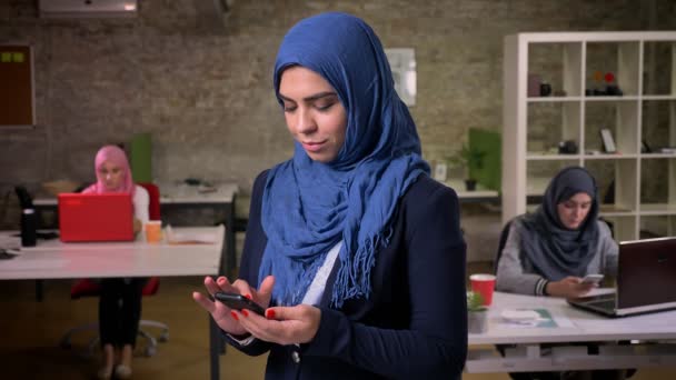 Awesome merokok perempuan arab dalam hijab biru gelap menggunakan smartphone sambil berdiri di kantor bata dekat gadis arab lain duduk di meja mereka, ilustrasi modern — Stok Video