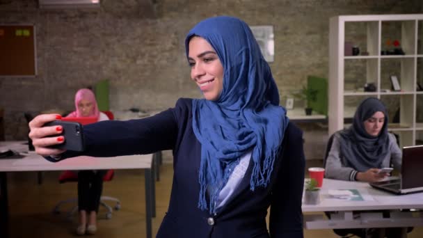 Насолоджуюча арабська жінка посміхається і приймає селфі в блакитному хіджабі і стоїть прямо орієнтовано на заморожування, в приміщенні, цегляний офіс — стокове відео
