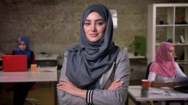 Muhteşem gri hijab sakin Arap kız gülümseyerek kameraya bakarak ve tuğla ofis yakınındaki diğer ayakta Ortadoğu kadın oturuyor