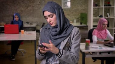 Ona telefon tutarak ve hijabs arka plan, çalışma ruh hali üzerinde kapalı, Müslüman kız ayakta iken swiping güzel Arap kız