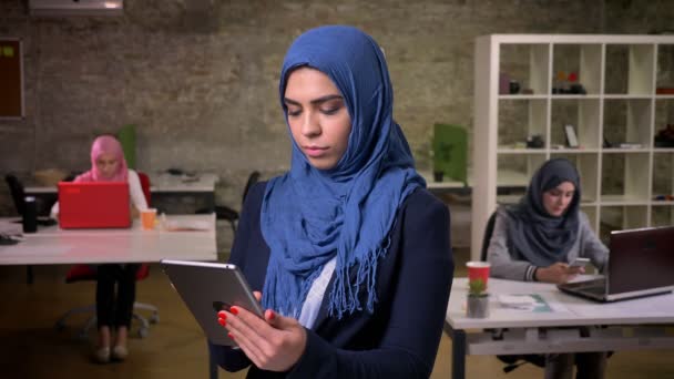 Serious terfokus arab perempuan di hijab biru berdiri dan mengetik di tablet nya, perempuan arab di latar belakang, modern ilustrasi di dalam ruangan — Stok Video