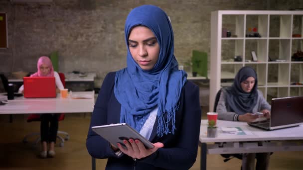 Senyum bahagia wanita arab yang menunjukkan layar hijau di tablet dan berdiri di kantor modern, wanita arab di latar belakang, bekerja getaran — Stok Video