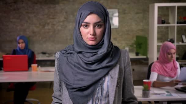 Mulher árabe bonita em hijab cinza está de pé calmamente e calmamente olhando para a câmera, fundo de tijolo, colegas de trabalho ar sentado e usando theis laptops — Vídeo de Stock
