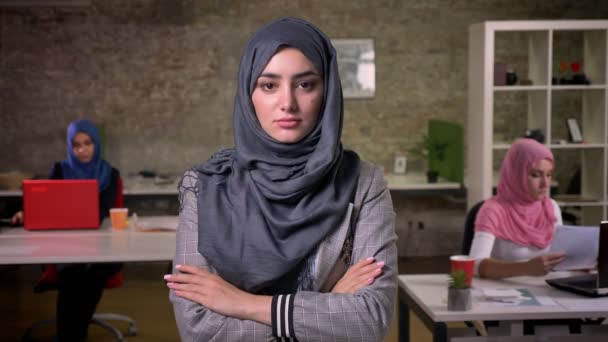 Ernstige aantrekkelijke Arabische vrouw staat voor het dragen van hijab rossed en camera kijken met rustig vertrouwen positie, baksteen studio, werkende meisjes achter — Stockvideo