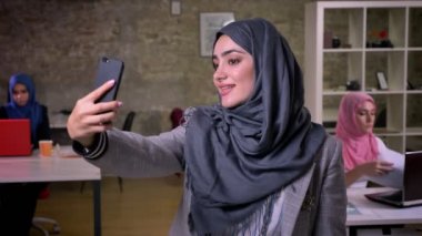 Tatlı gülümseyen Arap kadın onun smartphone kazandı kapalı duran, tuğla ışık ofis, onun arkasında çalışan kızlar selfies alıyor